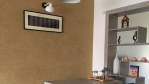 Un comedor con una mesa de concreto, dos sillas negras, un banco de madera, una lámpara colgante metálica y un estante con objetos decorativos.