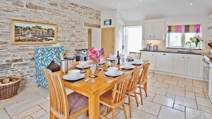 A dining area with a stone wall, a wooden table set for six, and a kitchen with white cabinets. - 4 bedroom accommodation in Mount, near Bodmin (Mount, near Bodmin)