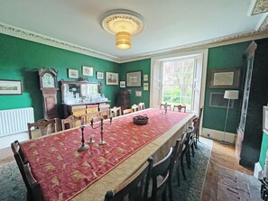 A dining room with a long table, chairs, a chandelier, a clock, and framed pictures on the wall.