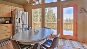 Une cuisine équipée d’un réfrigérateur en acier inoxydable, d’une table à manger avec des chaises et de grandes fenêtres donnant sur un paysage arboré et une terrasse.
