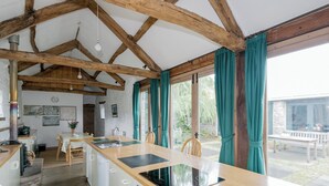 Een keuken met een plafond van houten balken, een centraal kookeiland met een spoelbak en uitzicht op de tuin door grote ramen.