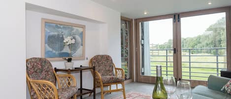 A room with two wicker chairs, a small table with a bottle of wine and a glass, and a large window offering a view of a green landscape.