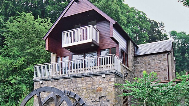 Una cabaña de madera con balcón y una noria grande adosada al costado.