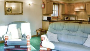 Sala de estar con dos sillones y un sofá, zona de cocina con armarios de madera, un microondas y una ventana con cortinas.