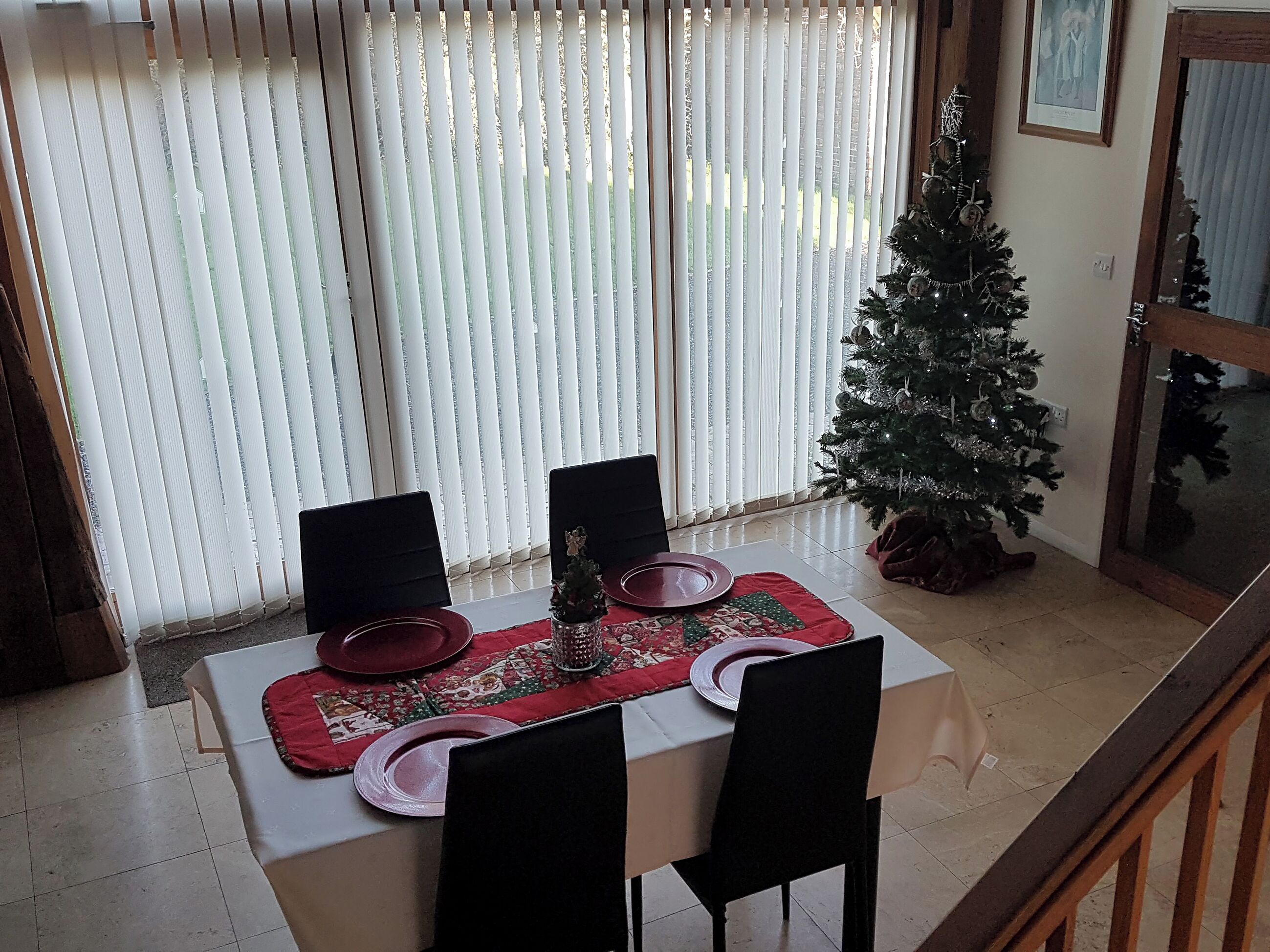 Un coin repas avec une table dressée pour deux personnes, un sapin de Noël et une fenêtre avec des stores.