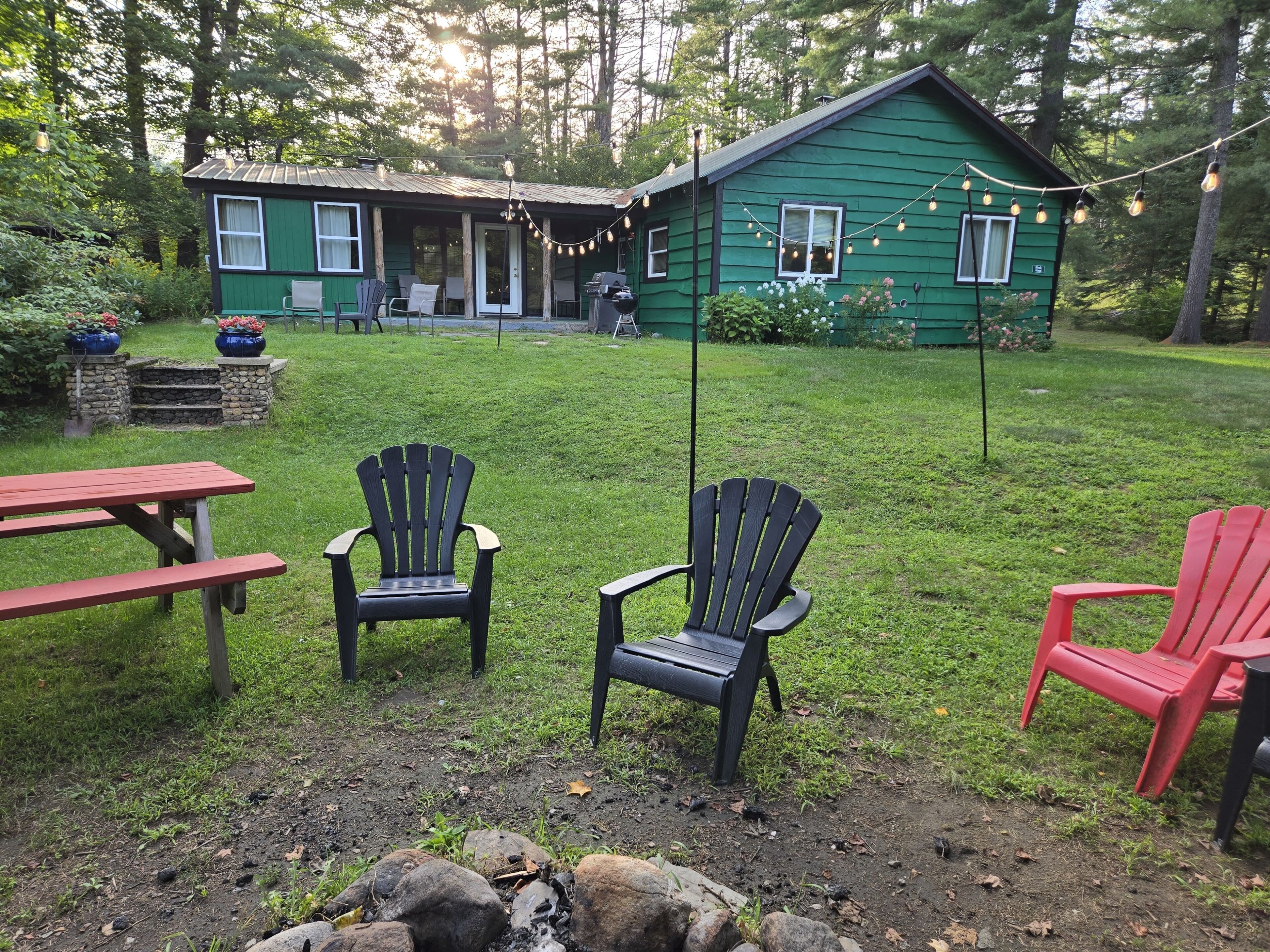 A green house with a porch, two Adirondack chairs, a red bench, and a picnic table.
