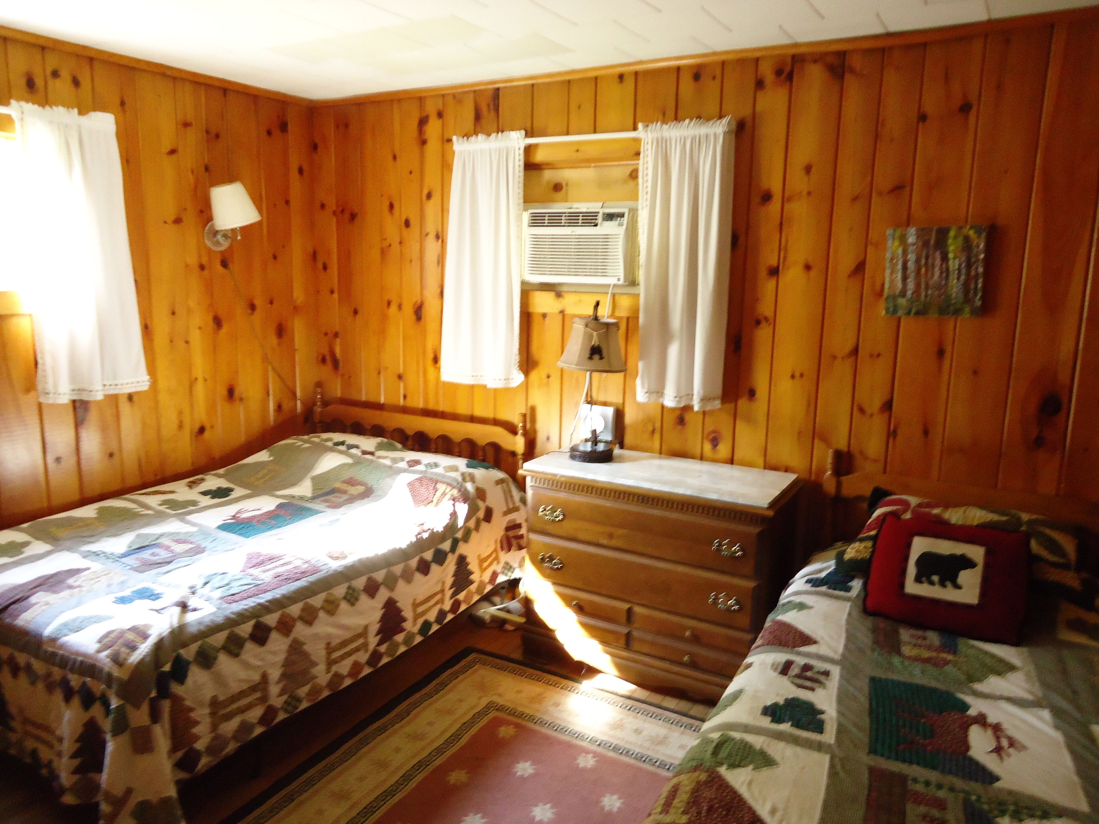 Una camera in stile baita con due letti, una cassettiera, una lampada e un condizionatore d'aria a parete.
