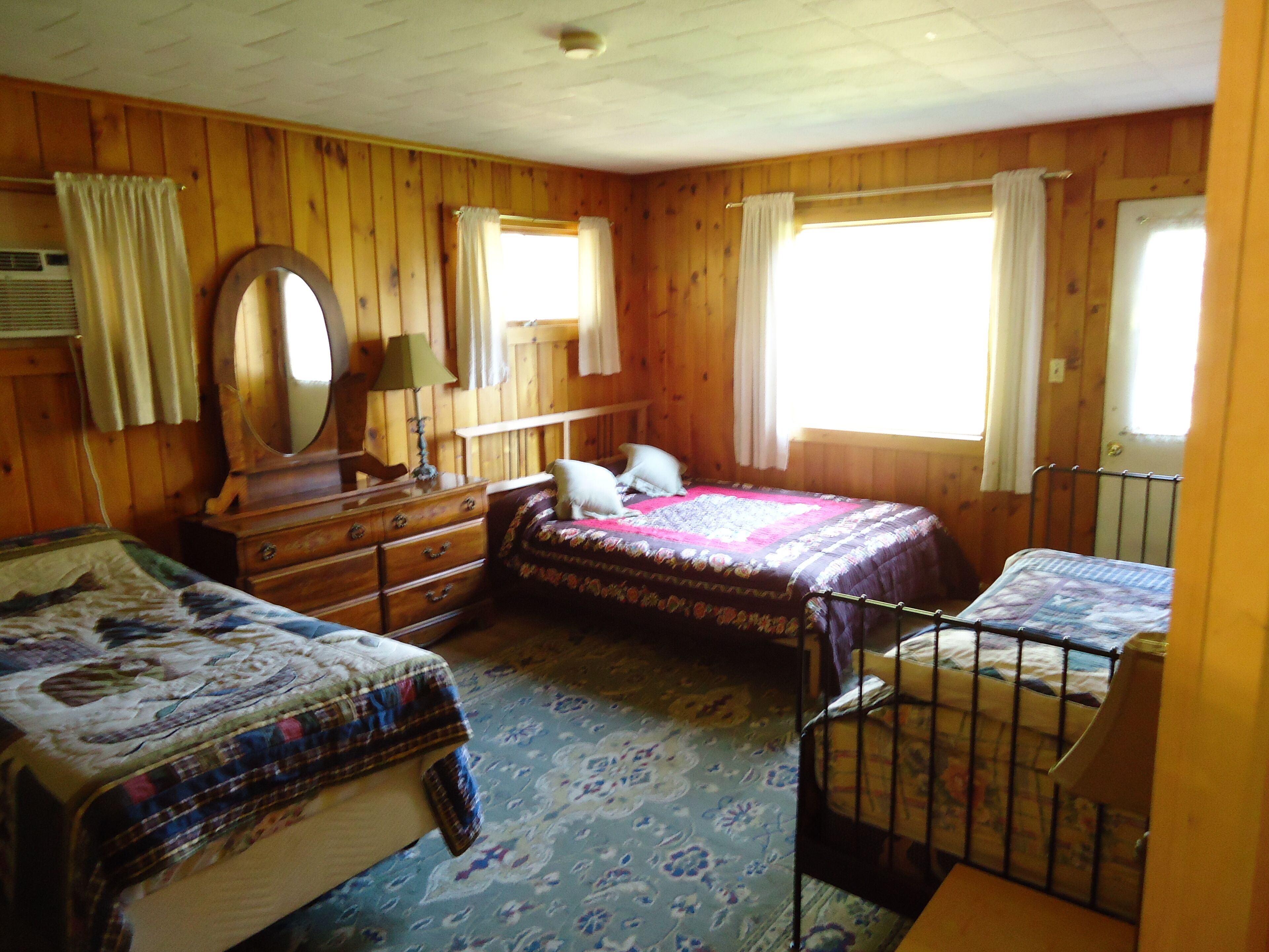 A wooden-paneled bedroom with two beds, a dresser, a mirror, and a window with curtains.