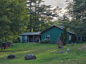 A green house with a porch, surrounded by a grassy yard and large rocks.