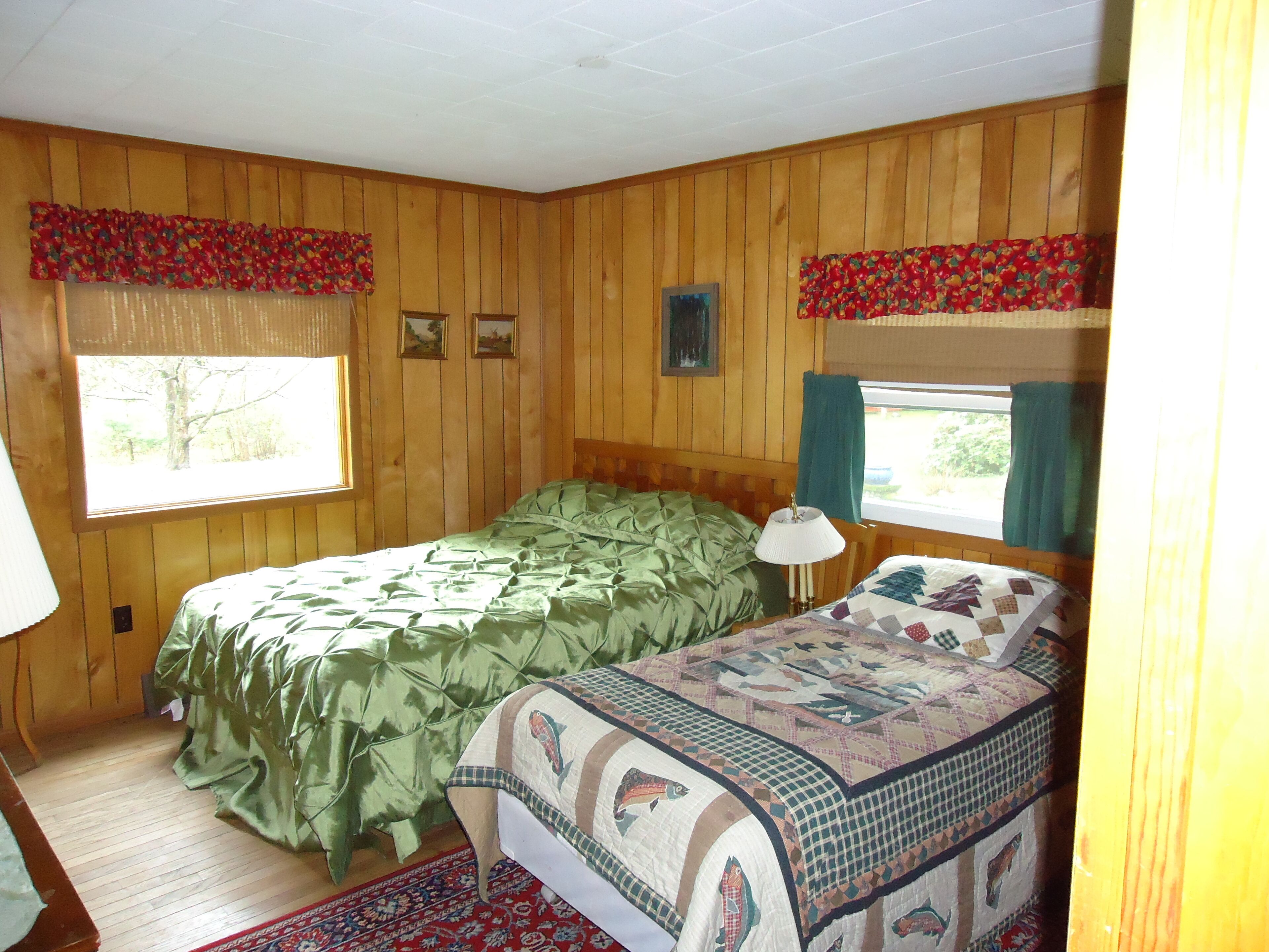 A room with two beds, wooden walls, a window with curtains, and a patterned rug.