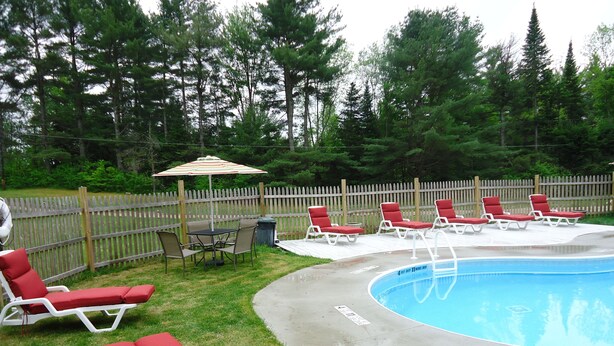 A backyard with a swimming pool, red lounge chairs, a table with chairs, and a wooden fence. - 5 Bedroom Pet Friendly House Lake George Region (Johnsburg)
