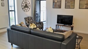 A modern living room with a black leather sofa, a television on a wooden stand, and a large window with a view of the outside. - 2 bedroom accommodation in Alnwick (Alnwick)