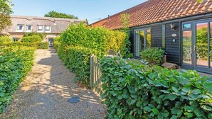 A narrow gravel path leading to a black building with a tiled roof, surrounded by lush greenery and a traditional house in the background. - Thyme for a relaxing break for two. (Catfield)