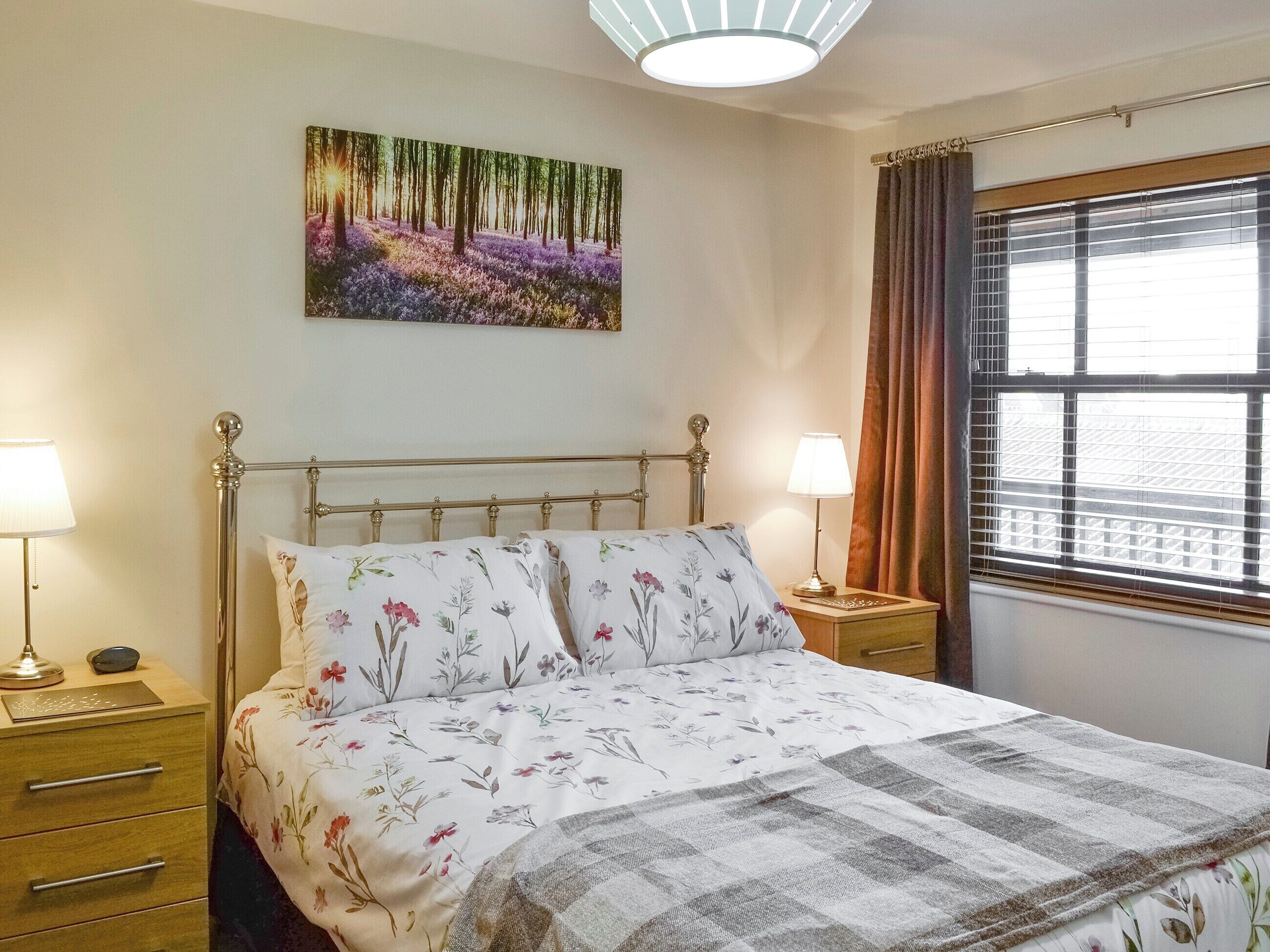A bedroom with a bed, two bedside tables, a nightstand, a lamp, a painting, a window with curtains, and a ceiling light.