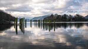 Een kalm meer met houten palen op de voorgrond, omringd door bomen en bergen in de verte.