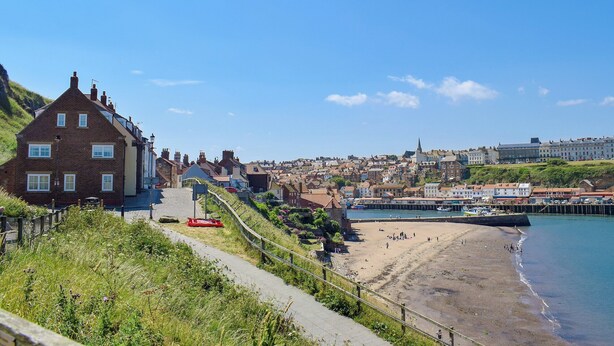 A coastal town with a prominent building on the left, a sandy beach, and a pier extending into the water. - Captains Cottage - E3643 (Whitby)