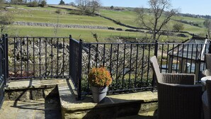 A balcony with a stone floor, a potted plant, wicker furniture, and a view of a green landscape with hills and a stone wall.