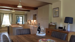A dining area with a wooden table, chairs, a wine bottle, and a glass. There is a kitchen in the background with cabinets and a window.