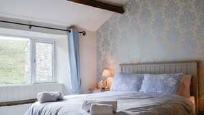 A bedroom with a stone wall, floral wallpaper, a wooden headboard, a window with a curtain, and a bedside table with a lamp.