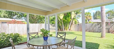 A covered patio with a round table and chairs, large windows, and a view of a backyard with a wooden fence and palm trees.