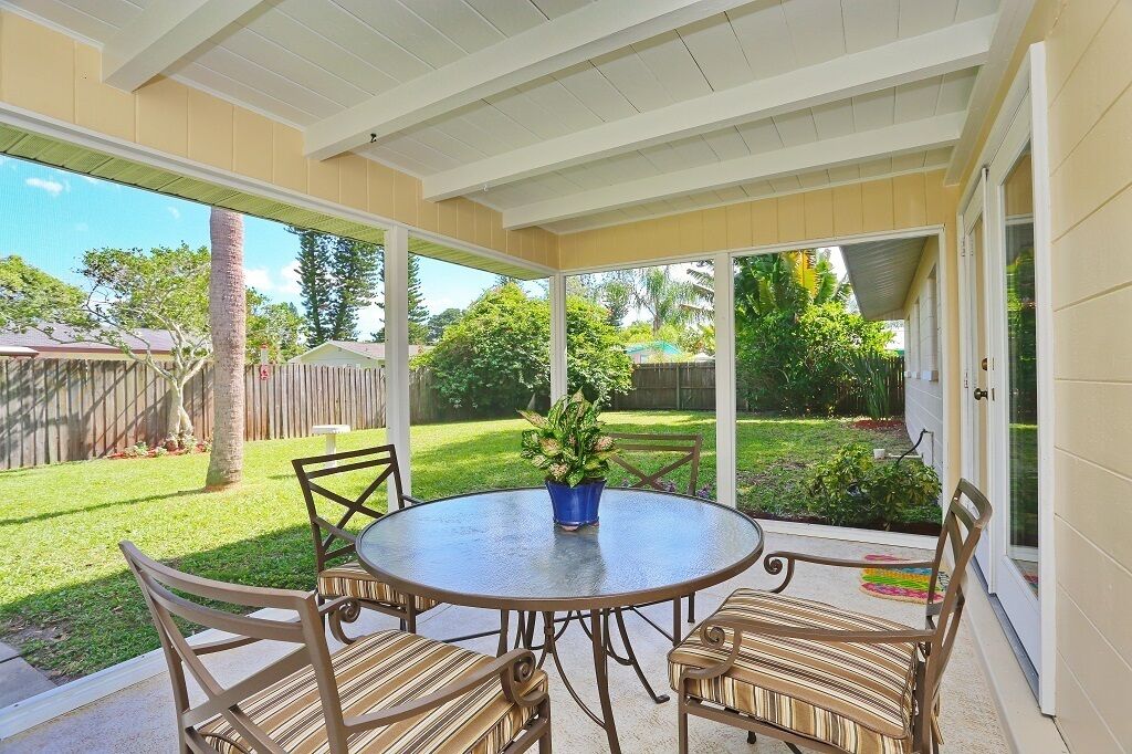 A covered patio with a round table and chairs, a potted plant, and a view of a backyard with a wooden fence and greenery.