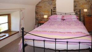 Une chambre avec un mur en pierre, un lit avec un cadre en métal, une commode en bois et une fenêtre avec des rideaux.