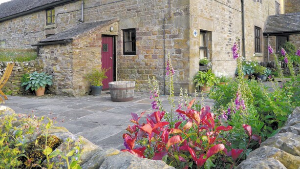 Un bâtiment en pierre avec une porte rouge, des plantes en pot et un jardin aux feuillages colorés.