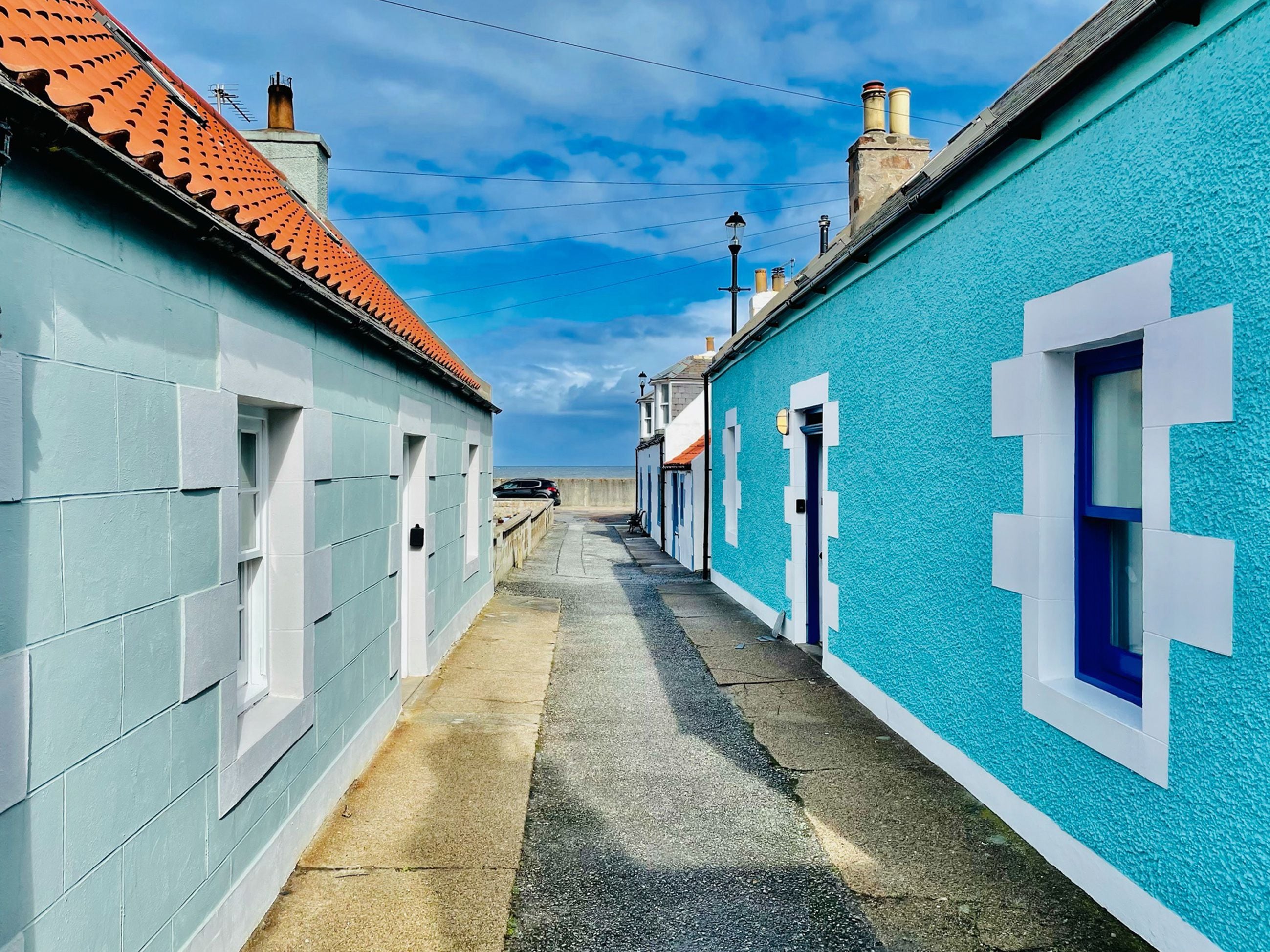 A narrow street with buildings on either side, featuring a mix of light and dark blue facades, a tiled roof, and a clear sky.
