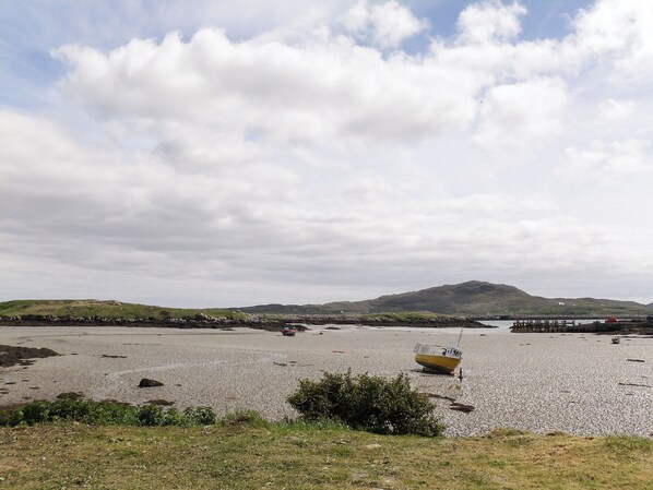 A coastal landscape with a small boat on a pebbly beach, a distant hill, and a pier.