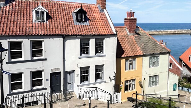 A coastal street with terracotta-tiled buildings, a white building with a black door, and a yellow building with a white fence.