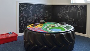 Una mesa redonda con cubierta a cuadros, un recipiente rojo con crayones y una pared de pizarra con dibujos.