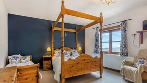 Una cama con dosel y tablero de madera, una mesita de noche con lámpara, una silla y una ventana con cortinas.