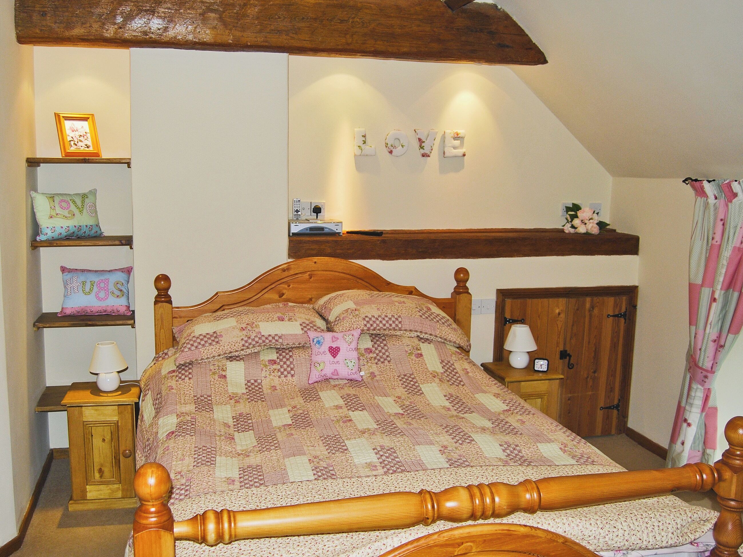 A bedroom with a wooden bed, a quilt, a shelf with decorative pillows, a wall-mounted clock, and a 'LOVE' sign.