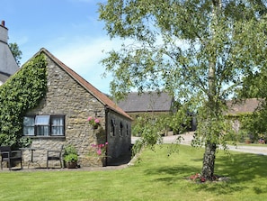 A stone building with a sloped roof, a small porch with chairs, and a tree in the foreground.