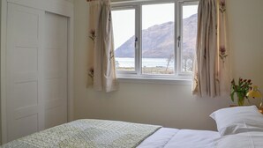 A bedroom with a bed, a window with curtains, a view of a mountain and water, and a bedside table with a lamp and flowers. - 4 bedroom accommodation in Taynuilt, near Oban (Taynuilt, near Oban)