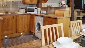 A compact kitchen with wooden cabinets, a microwave, a washing machine, and a dining area with a table set for two. - 1 bedroom accommodation in Watermillock-on-Ullswater (Watermillock-on-Ullswater)