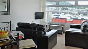 Ein Wohnzimmer mit Blick auf einen Jachthafen, ein schwarzes Ledersofa, ein kleiner Tisch mit Obstschale und ein an der Wand befestigter Fernseher.