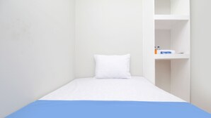 A small, single-bed room with a white wall and a blue bedspread. There is a white pillow on the bed. A white shelf unit is visible in the background. - Kamar Keluarga Bandara Mas Syariah (Tangerang)