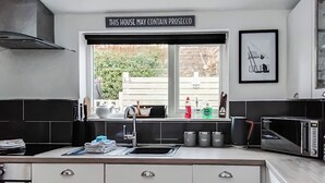 Eine moderne Küche mit Schwarz-Weiß-Farbschema, ein Fenster mit Blick auf Grünflächen und ein Schild über dem Fenster mit der Aufschrift 'THIS HOUSE MAY CONTAIN PROSECCO'.