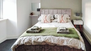 Ein ordentlich gemachtes Bett mit grüner Decke und zwei gefalteten Handtüchern, ein Nachttisch mit Lampe, eine Wandablage und ein Spiegel.