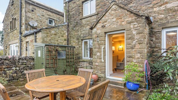 A stone house with a wooden patio table and chairs, a green gate, and a garden hose.