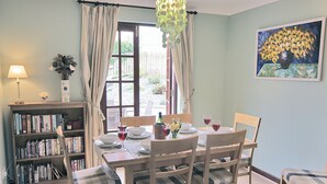 A dining room with a table set for four, a bookshelf, a painting, and a window with curtains. - Pebble Cottage - HW7447 (Broad Haven)