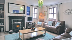 A living room with a fireplace, a television, a sofa, a coffee table, and a chandelier. - Pebble Cottage - HW7447 (Broad Haven)
