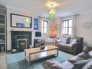 A living room with a fireplace, a television, a sofa, a coffee table, and a chandelier. - Pebble Cottage - HW7447 (Broad Haven)