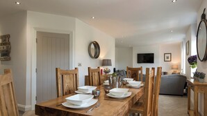 Un coin repas avec une table en bois dressée pour quatre personnes, entourée de chaises en bois. Un téléviseur est fixé au mur, et un canapé se trouve au fond de la pièce.
