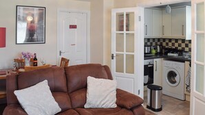 Ein Wohnzimmer mit braunem Sofa, einem Esstisch mit Stühlen, einer Küche mit Geräten und einer Tür mit einem Schild.