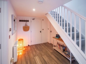 A hallway with a wooden floor, a white door, a staircase with white railings, and a shelf with books and a bag. - Red Squirrel Cottage (Whitwell, near Ventnor)