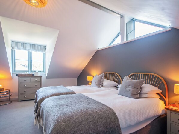A bedroom with a sloped ceiling, a bed with pillows, bedside lamps, a dresser, and a window with curtains. - Red Squirrel Cottage (Whitwell, near Ventnor)