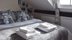 Een slaapkamer met een bed, kussens, een radiator, een raam met gordijnen en lijstjes met afbeeldingen aan de muur.