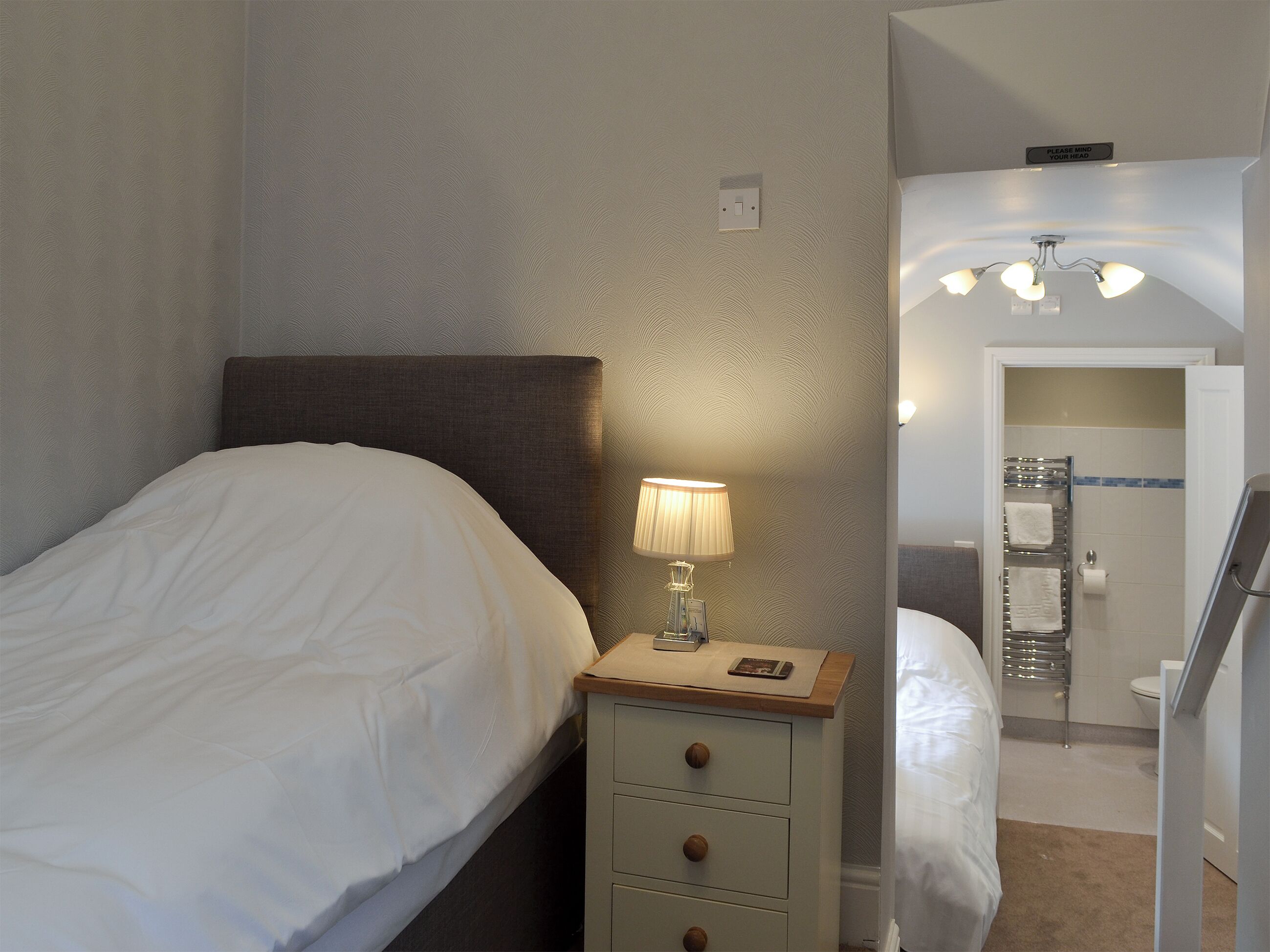 A neatly made bed with white linens, a bedside lamp, and a small wooden nightstand with drawers.